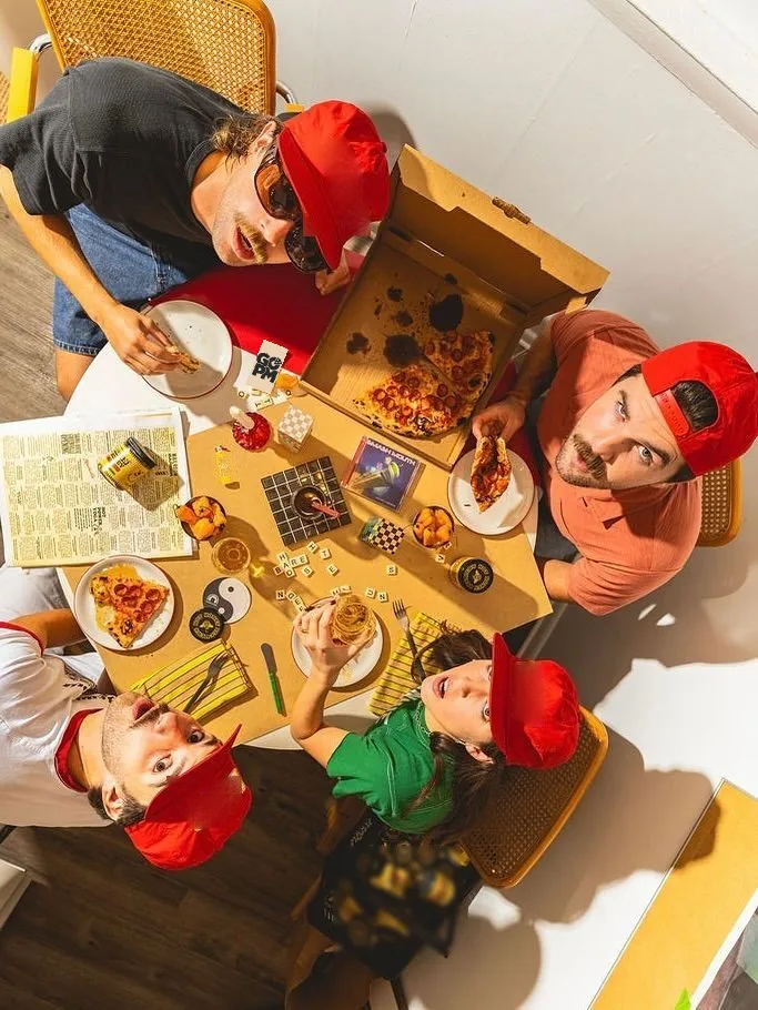 Vogelperspektive: vier Freunde in roten GO-PM-Caps um einen Tisch herum, eine geöffnete GO-PM-Pizzaschachtel mit Pepperoni, Getränke, Snacks und ein Brettspiel-Setup.