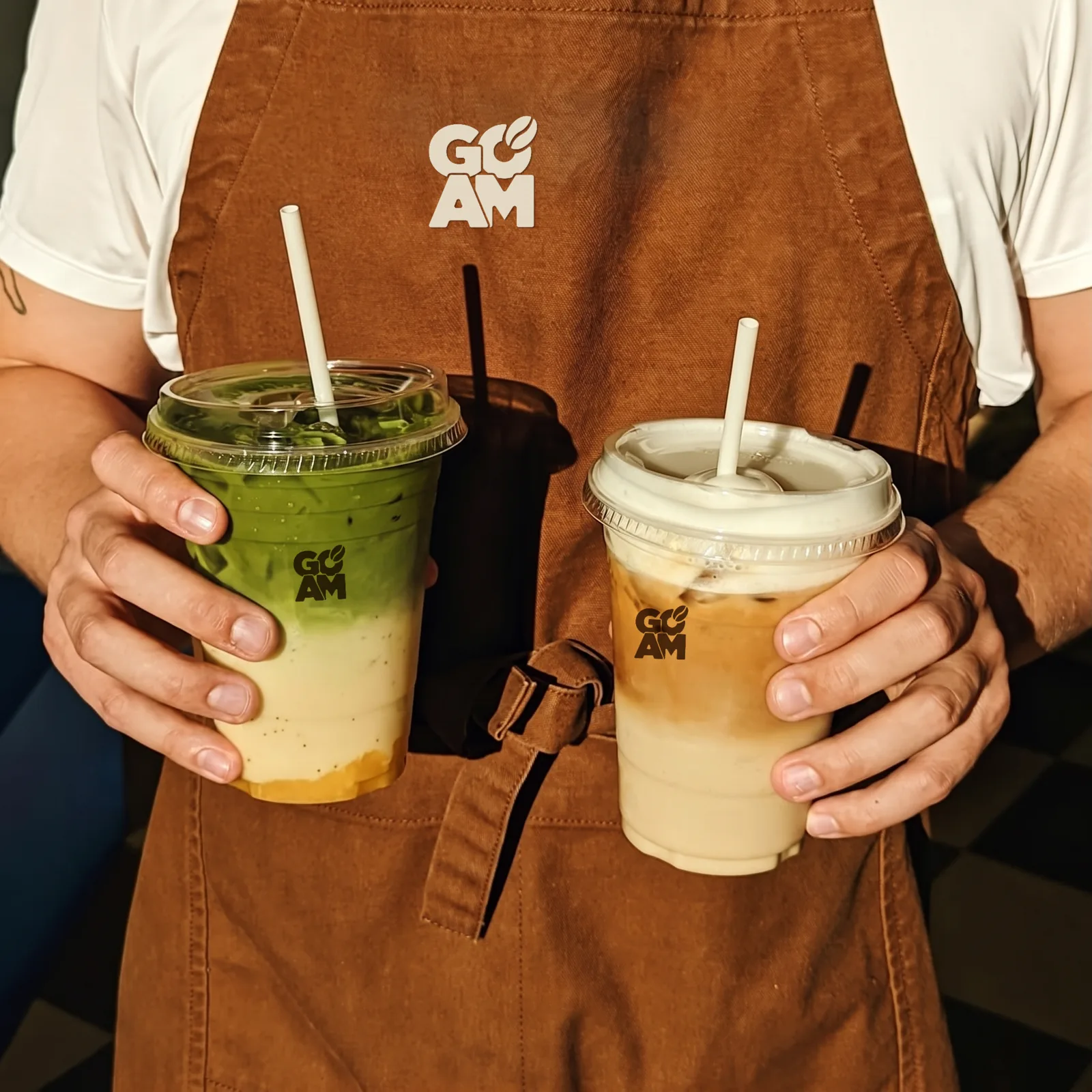 Barista in karamellbrauner GO AM Schürze hält zwei gebrandete Becher — Matcha-Getränk und Iced Latte.