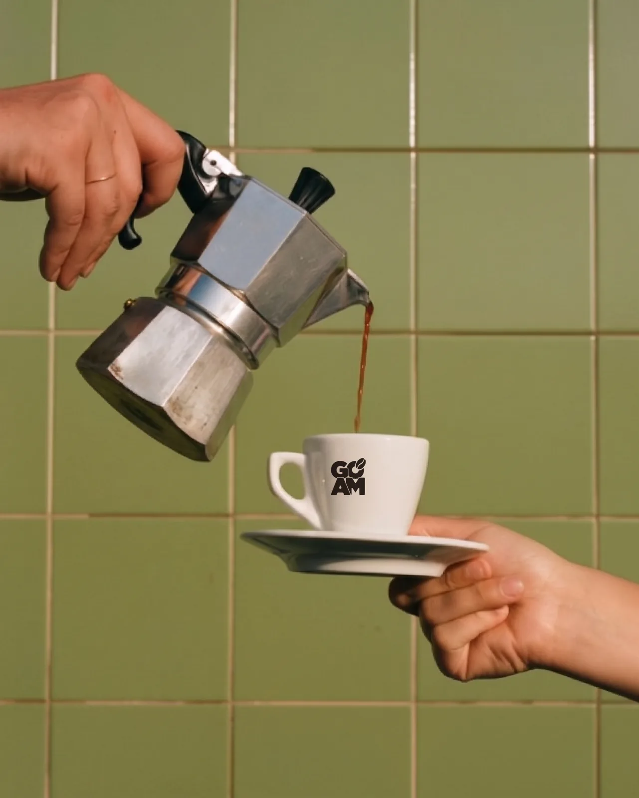 Eine Hand giesst Espresso aus einer Bialetti-Moka-Kanne in eine weisse GO PM Espresso-Tasse, die eine zweite Hand entgegenhält, vor salbeigrüner Kachelwand.