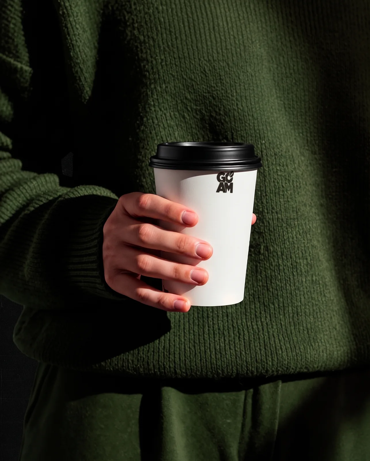 Hand in dunkelgrünem Strickpulli hält einen gebrandeten GO PM To-Go-Becher mit schwarz-weissem Checker-Band und Bean-Lockup.